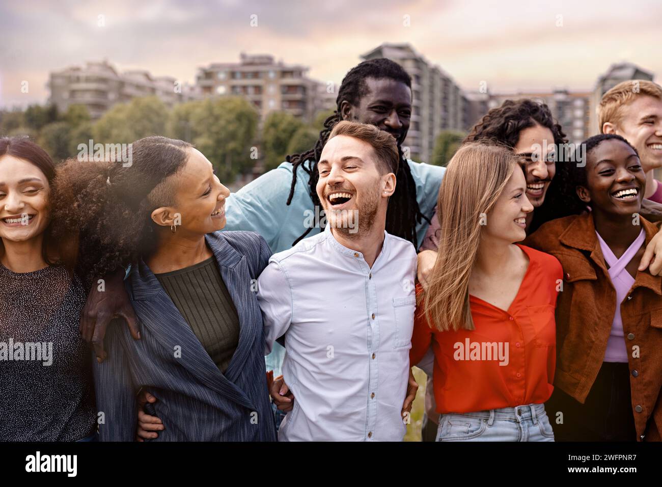 Eine überschwängliche Gruppe junger Erwachsener aus verschiedenen ethnischen Hintergründen, die einen Moment des echten Lachens und Genusses vor einer urbanen Kulisse teilen - Beau Stockfoto