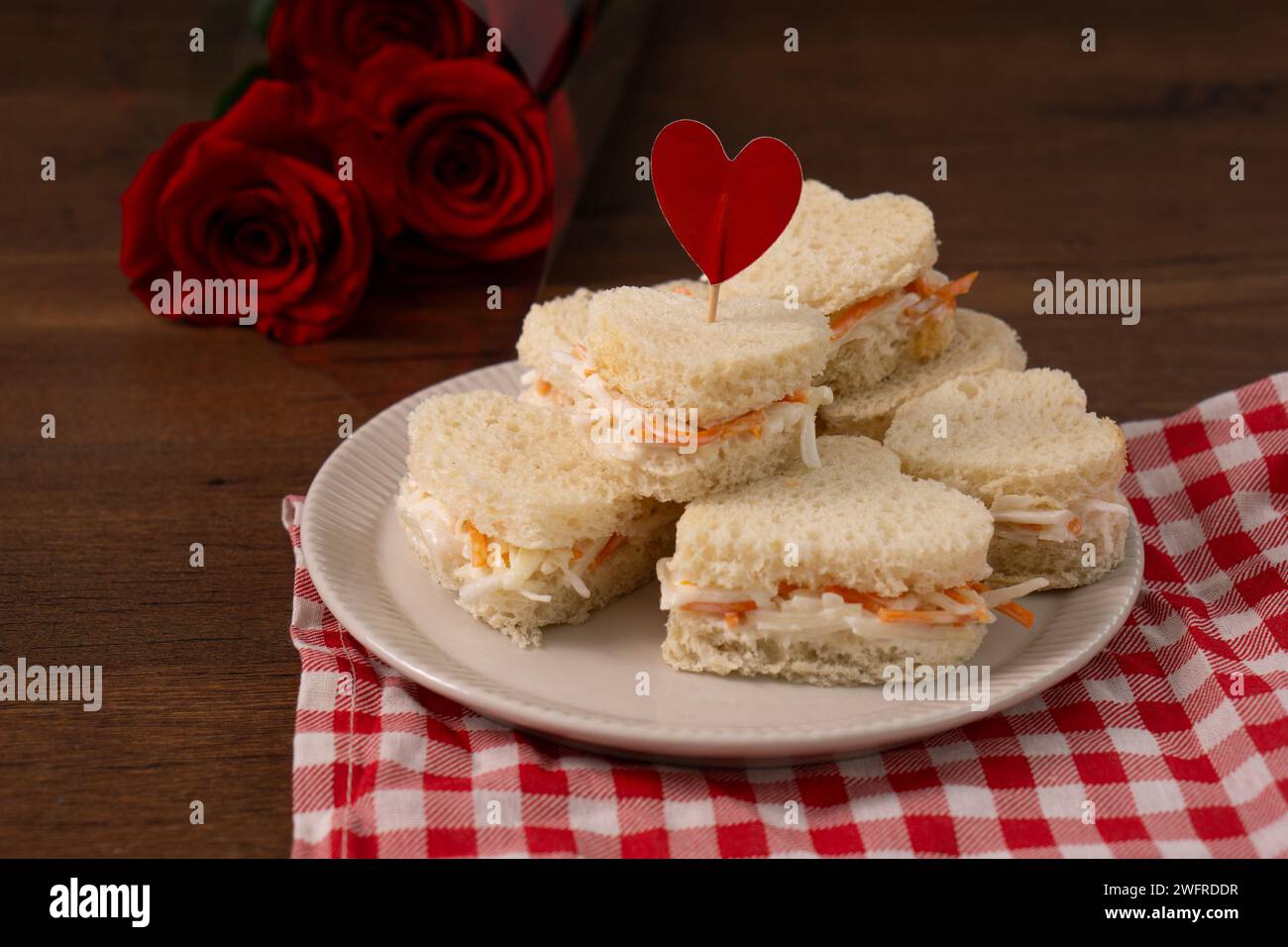 Herzförmige Sandwiches aus Salat, die auf einer weißen Schale auf einem rot-weiß karierten Tuch liegen. Auf dem Hintergrund steht ein Rosenstrauß. st. Va Stockfoto