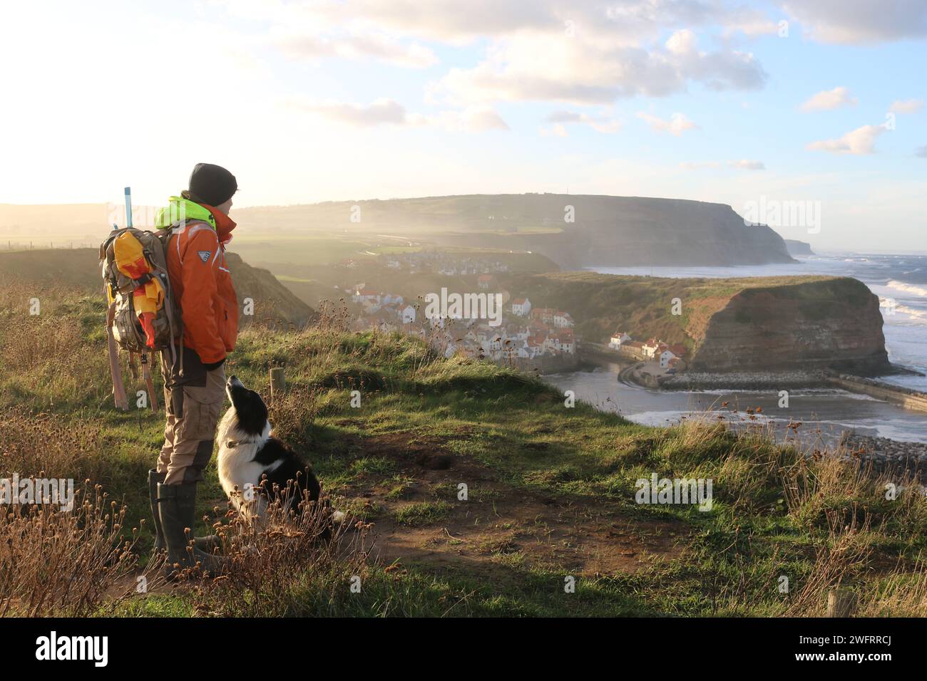 Ein Mann und sein Border Collie Hund, auf dem Cleveland Way Pfad, oberhalb des alten Fischerdorfes Staithes während eines trüben Sonnenuntergangs. Stockfoto