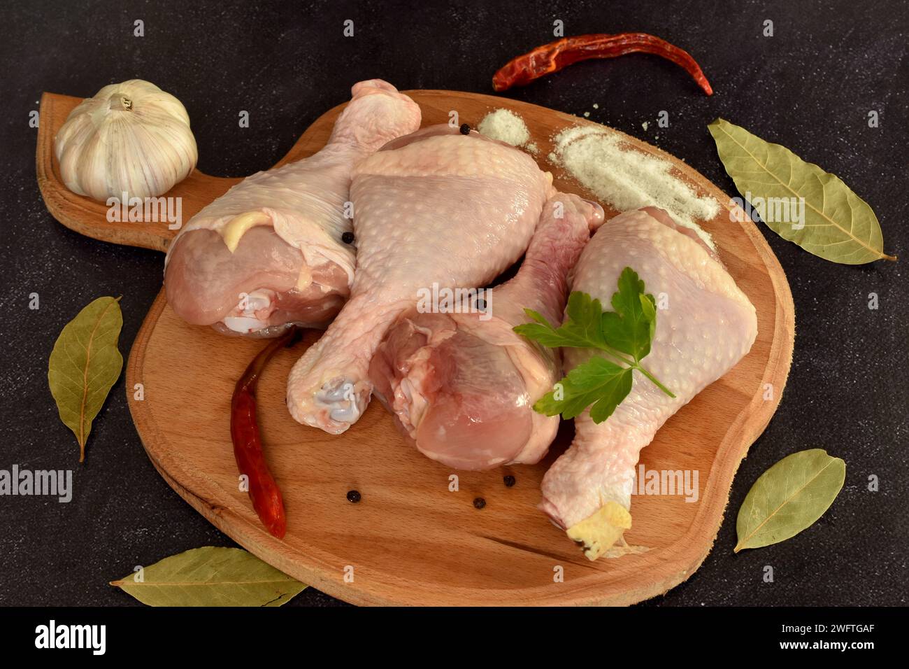 Auf einem dunklen Hintergrund, auf einem Brett, liegen rohe Hähnchenschenkel bereit zum Kochen. Neben dem Fleisch befinden sich Lorbeerblätter, Pfeffer und Knoblauch. Stockfoto