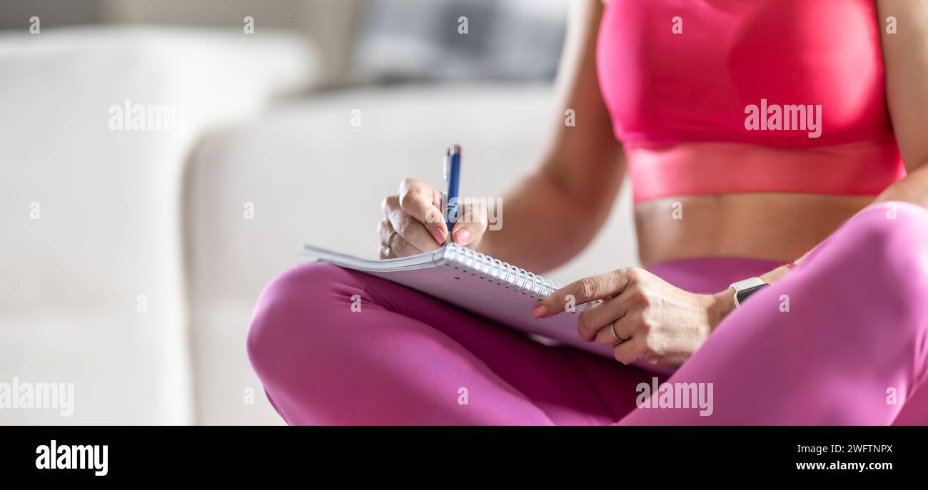 Sportliche Frau sitzt auf einer Matte und schreibt zukünftige Trainingspläne auf, um sportliche Ergebnisse zu erzielen. Stockfoto