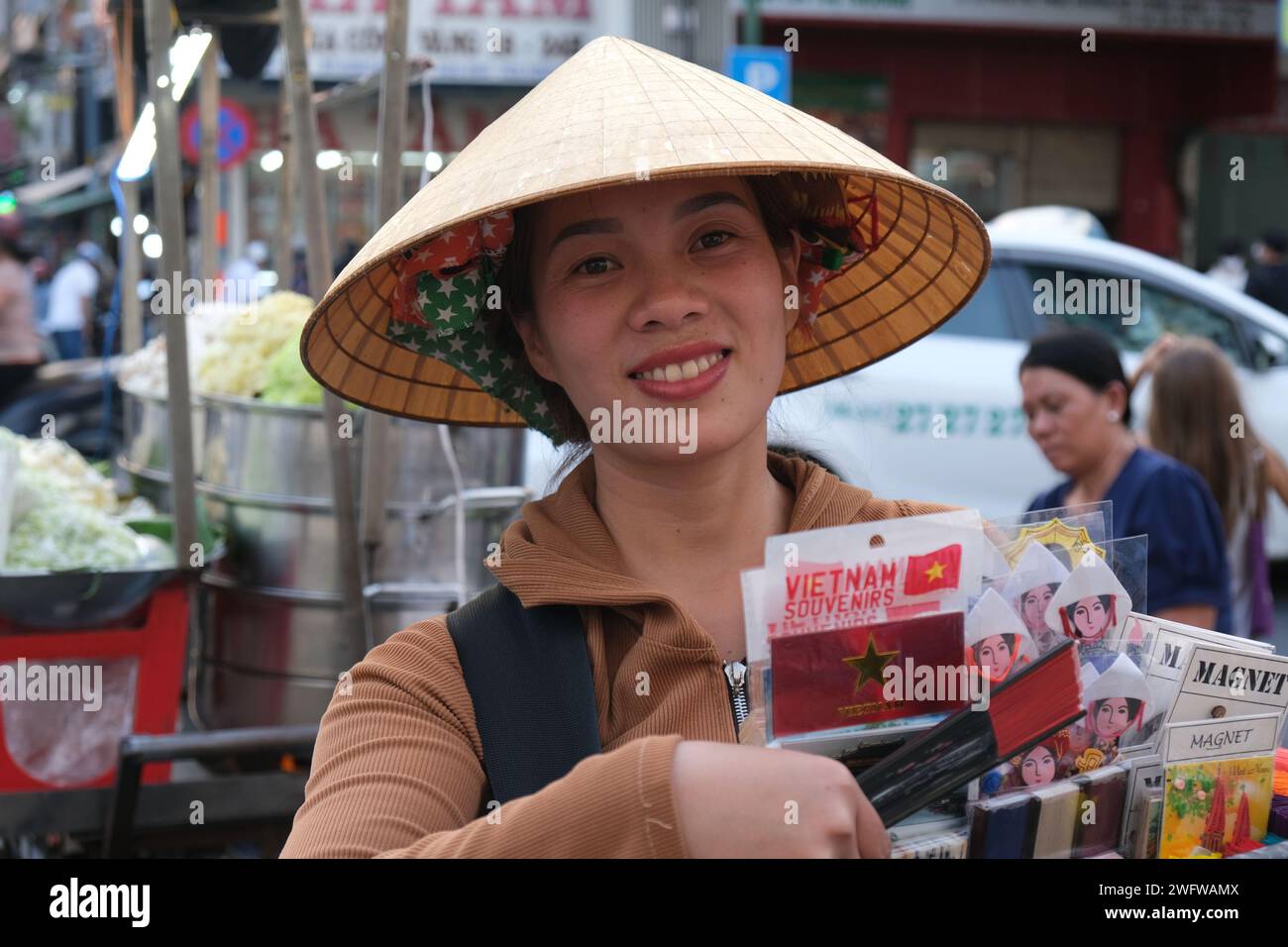 Lächelnde Frau mit konischem Hut, die Souvenirs auf der Straße vor einem geschäftigen Markt in Ho-Chi-Minh-Stadt, Vietnam, verkauft Stockfoto