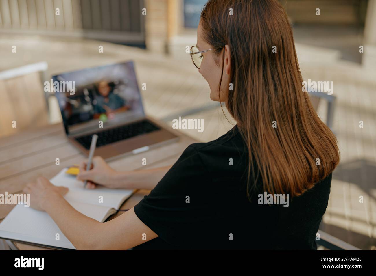 Geschäftsfrau, die mit dem Kunden per Videoanruf spricht und Notizen macht, während sie auf der Terrasse des Cafés sitzt Stockfoto
