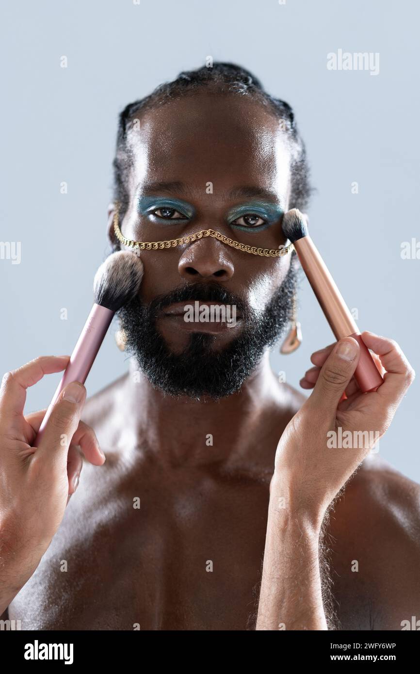 Porträt eines afroamerikanischen schwulen Mannes, der Make-up mit Pinseln aufträgt, die von Crop Artist gehalten werden. Nahaufnahme eines homosexuellen Mannes mit hellem Lidschatten und goldener Kette, der in die Kamera auf blauem Hintergrund blickt. Stockfoto