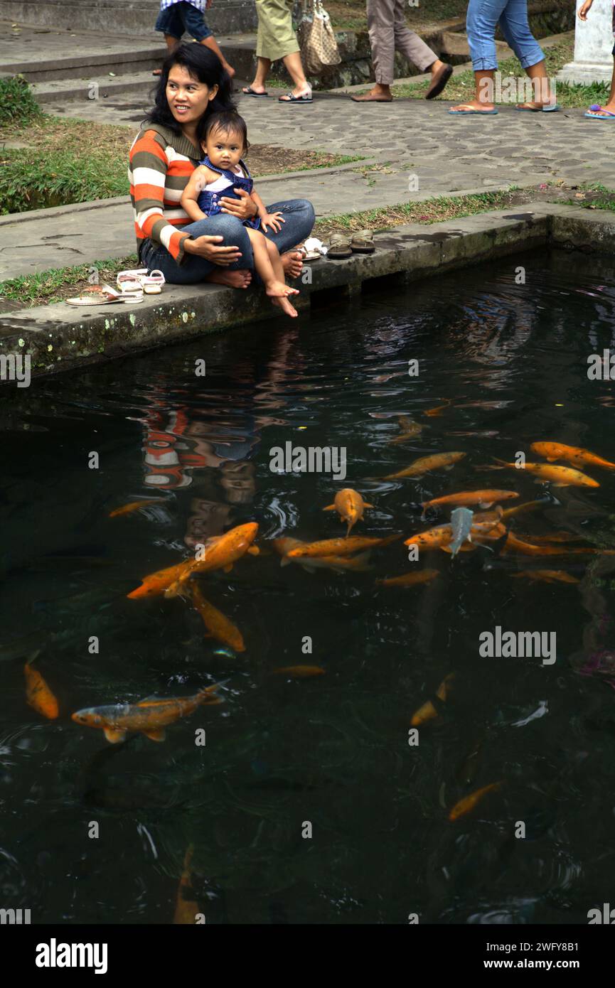 Eine Frau kümmert sich um ein Baby, während sie auf der Seite eines Teichs mit Goldfischen im Tirta Gangga Wasserpalast sitzt, einem ehemaligen königlichen Palast, der heute eine beliebte Touristenattraktion ist, befindet sich in Karangasem, Bali, Indonesien. Stockfoto