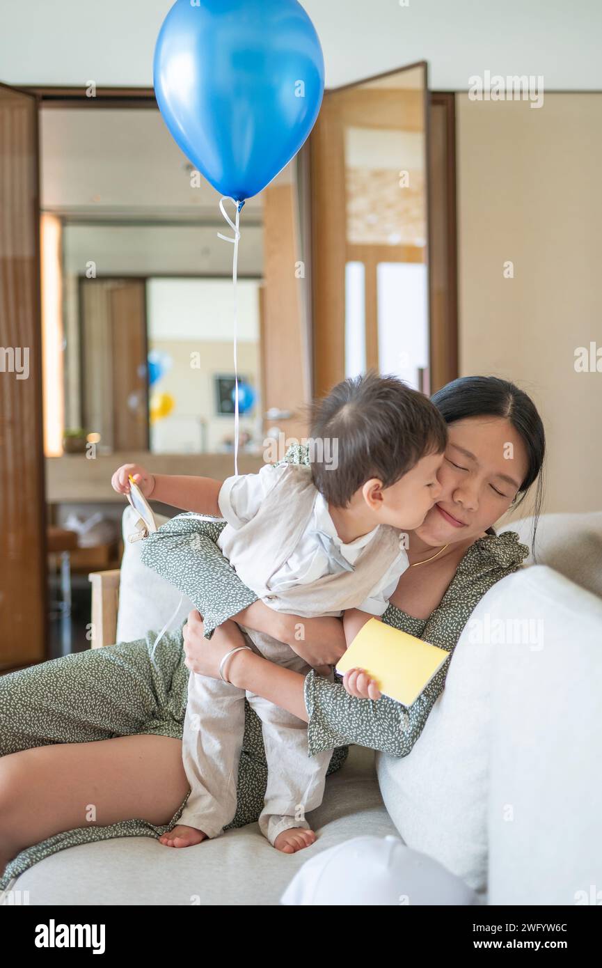 Niedlicher, einjähriger, multirassischer Junge, der seine Mutter küsst, während er seinen ersten Geburtstag mit seiner Mutter feiert und zu Hause einen blauen Ballon trägt Stockfoto
