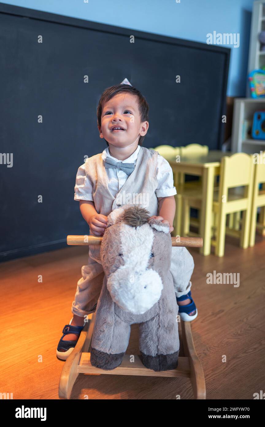 Niedlicher einjähriger multirassischer Junge, der seinen ersten Geburtstag im Kindergarten feiert. Er reitet auf einem hölzernen Spielzeugpferd und lächelt süß Stockfoto