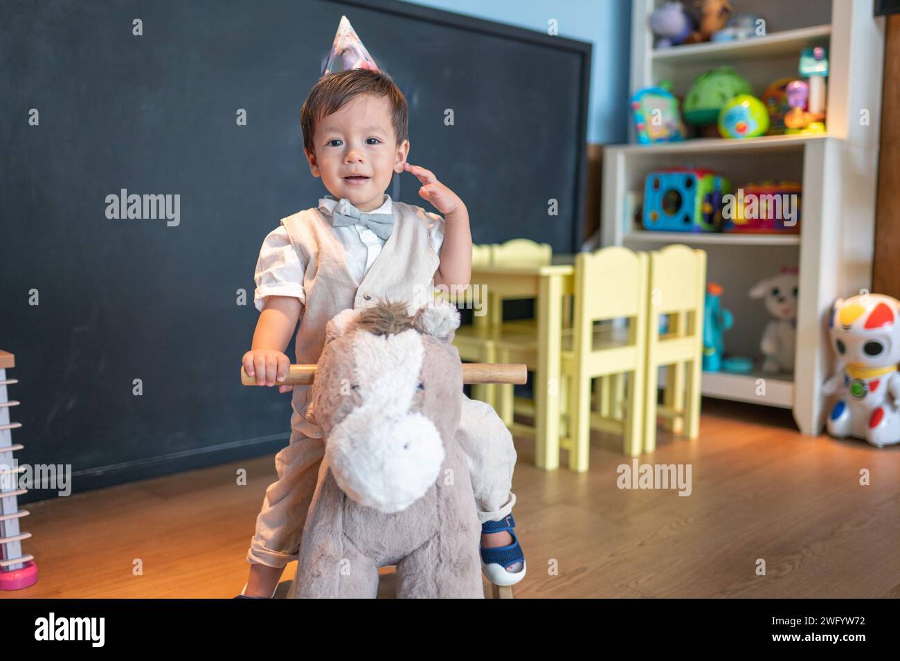 Niedlicher einjähriger multirassischer Junge, der seinen ersten Geburtstag im Kindergarten feiert. Er reitet auf einem hölzernen Spielzeugpferd und lächelt süß Stockfoto
