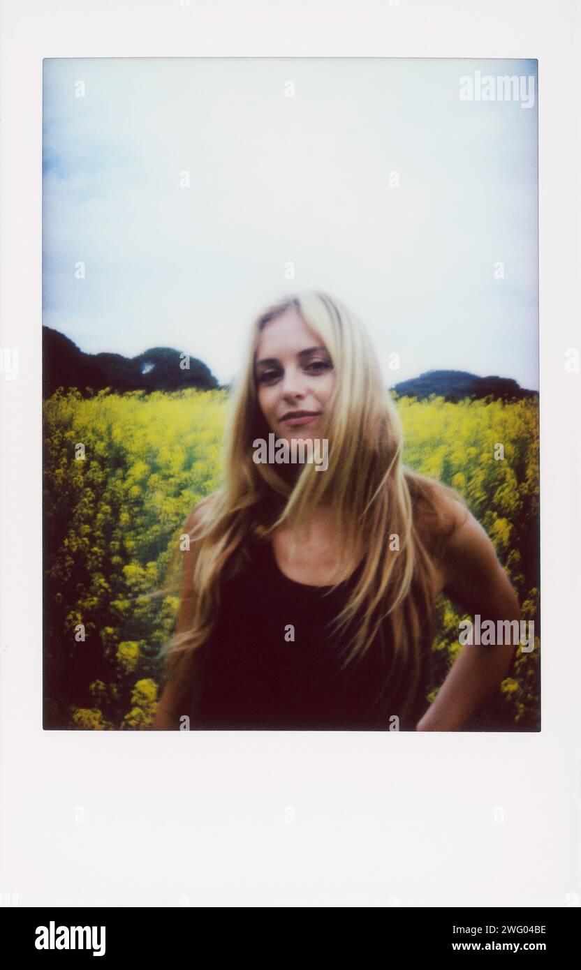 Sofortiges Bild einer schönen jungen Frau, die zwischen gelben Blumen steht. Junge blonde Frau auf einem Rapsfeld zur Frühlingszeit. Stockfoto