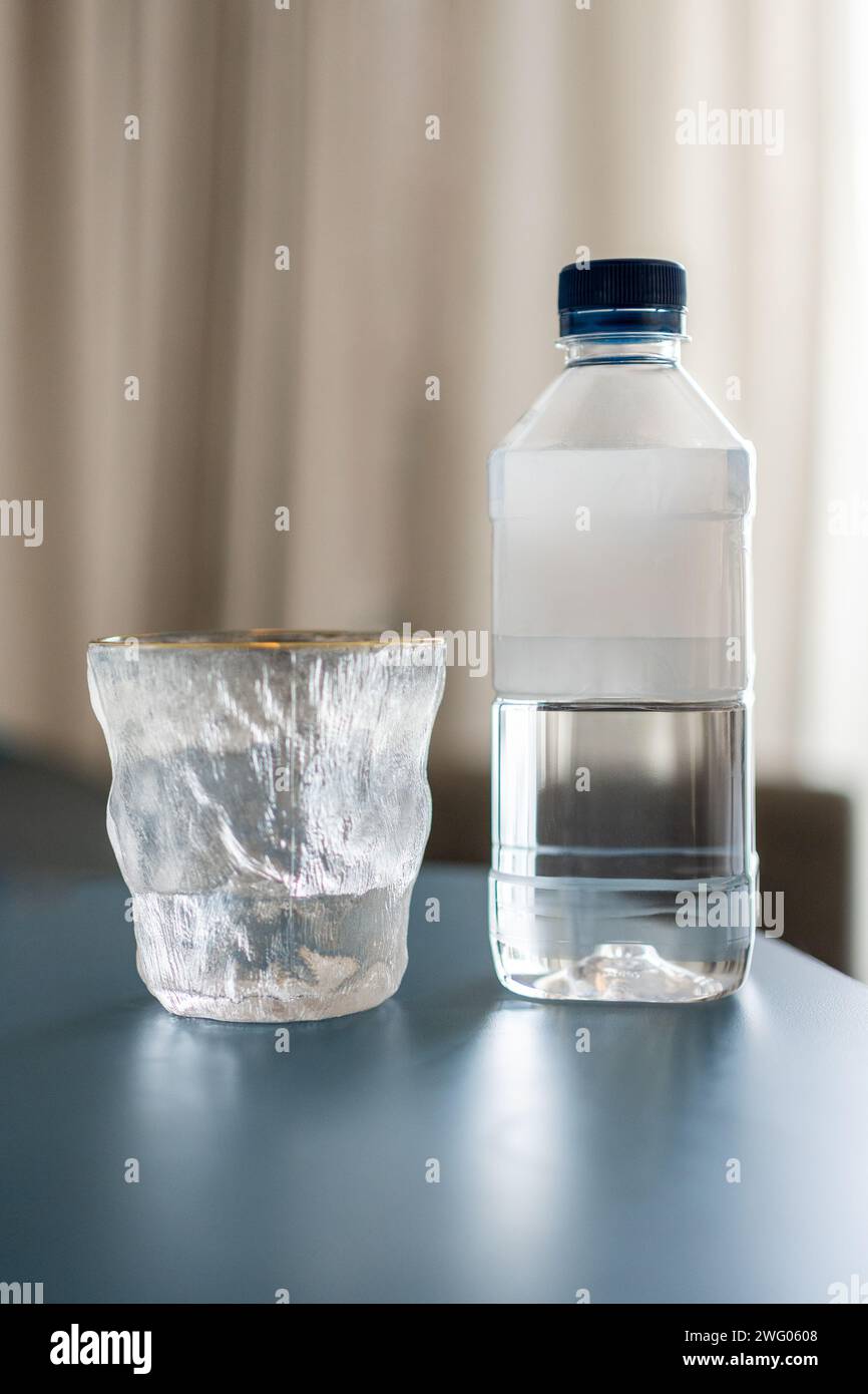 Trinkwasserflasche aus Kunststoff, die auf einem blaugrünen Tisch neben einem Designerglas mit unscharfem Hintergrund sitzt, Platz für Kopierraum oben und unten Stockfoto