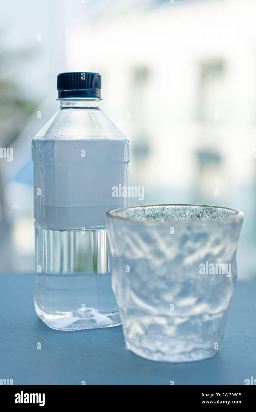 Trinkwasserflasche aus Kunststoff, die auf einem blaugrünen Tisch neben einem Designerglas mit unscharfem Hintergrund sitzt, Platz für Kopierraum oben und unten Stockfoto