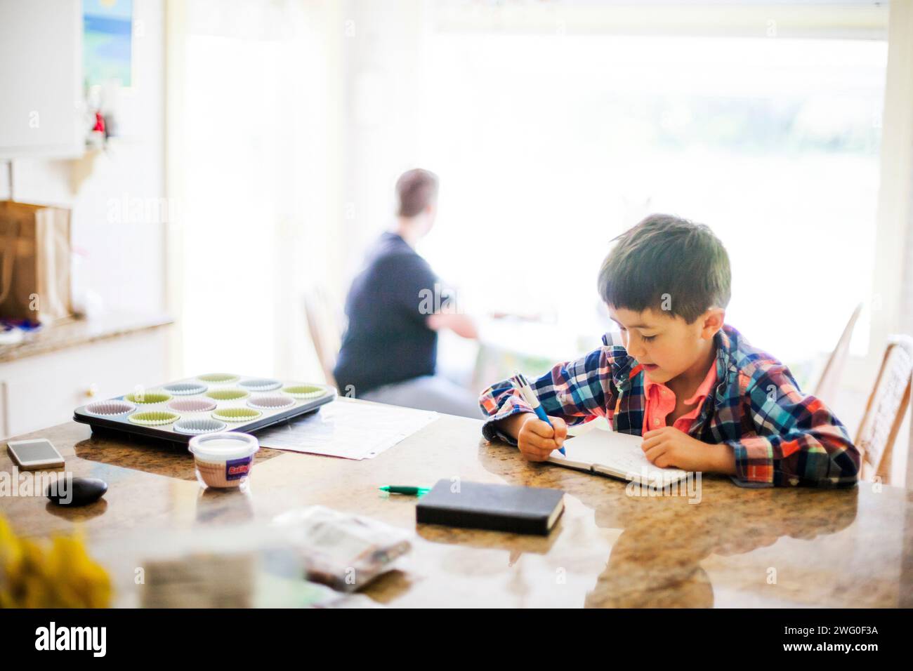 Ein japanischer amerikanischer 6-jähriger Junge schreibt in seinem Tagebuch an der Küchentheke. Hinter ihm sitzt ein Mann in einer Küchenecke. Stockfoto