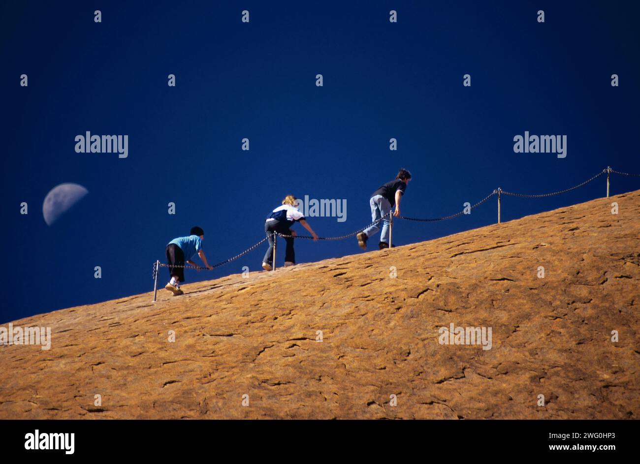 Drei Menschen besteigen den Uluru im Northern Territory Australiens, unterstützt von den Ketten und unterstützt von einem Halbmond. Stockfoto