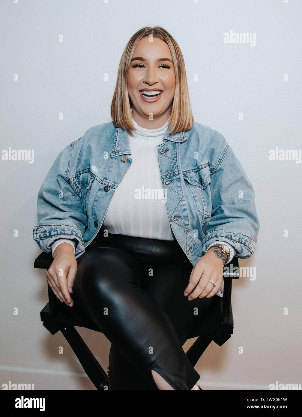 Das kurze blonde Mädchen in jeansjacke sitzt und wartet an der weißen Wand Stockfoto
