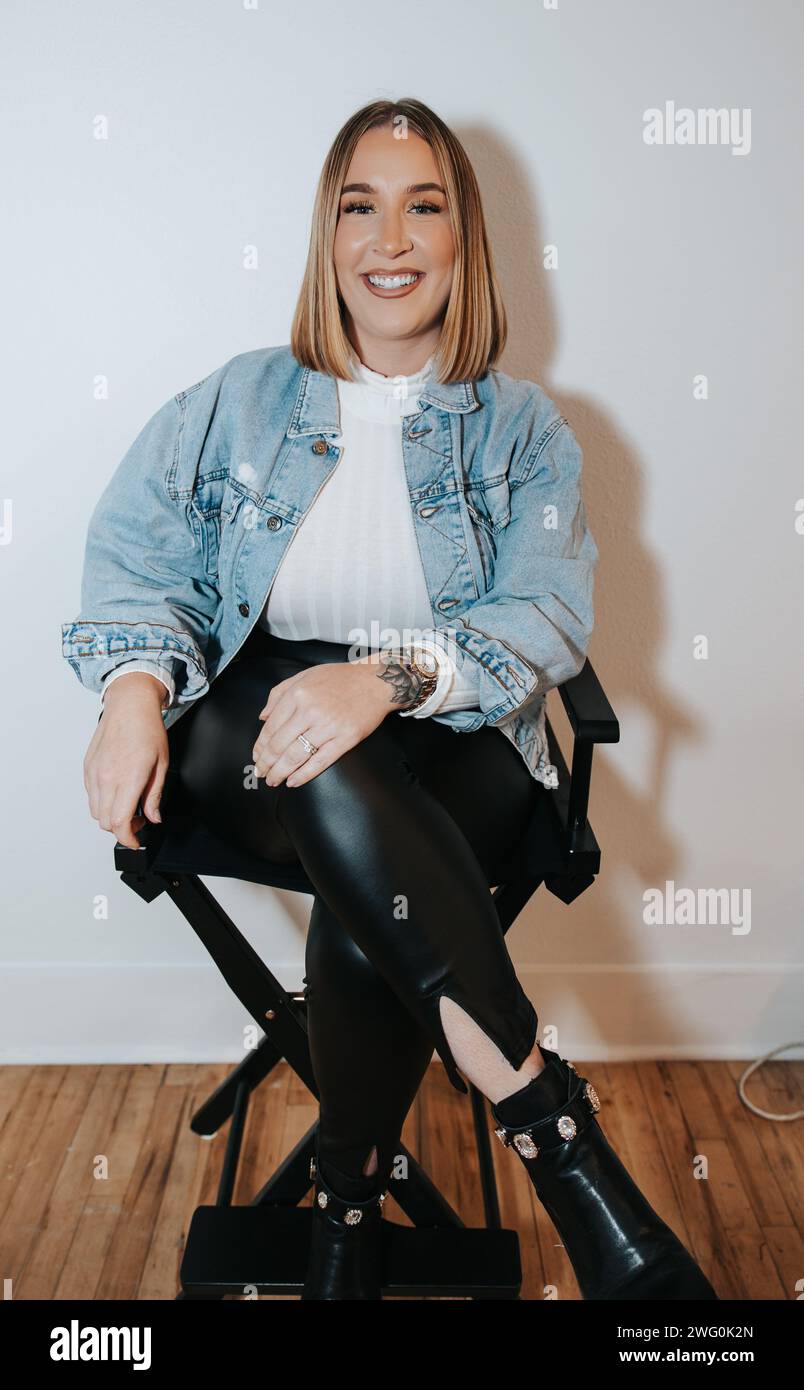 Das kurze blonde Mädchen in jeansjacke sitzt und wartet an der weißen Wand Stockfoto