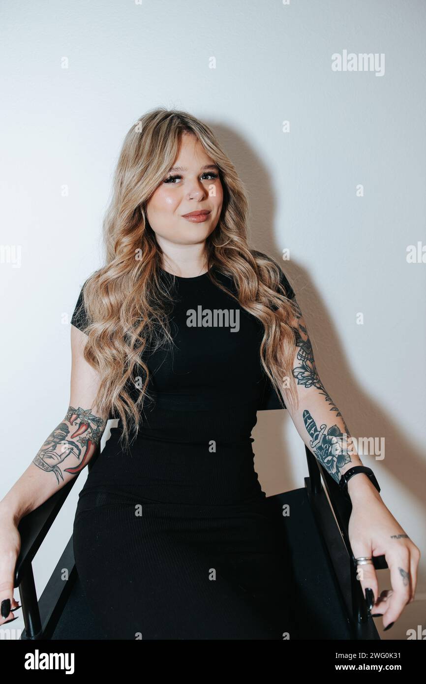 Blondes Mädchen in schwarzem Kleid sitzt und wartet an einer weißen Wand Stockfoto