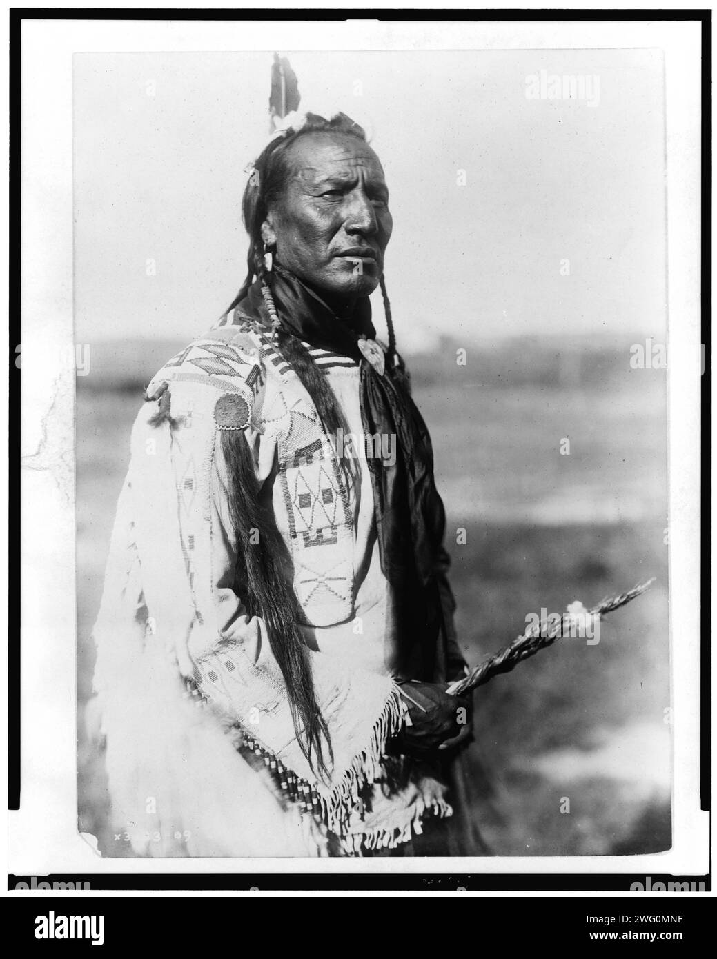 Große Mundfeder, c1910. Big-Mouth-Feder, Dreiviertelformat, stehend, nach rechts gerichtet, Zöpfe, eine Feder, ein perlenbesetztes Hemd aus Buchsleder, ein verziertes Kopfhautschloss auf der rechten Schulter, ein schwarzes Seidenhalstuch. Stockfoto