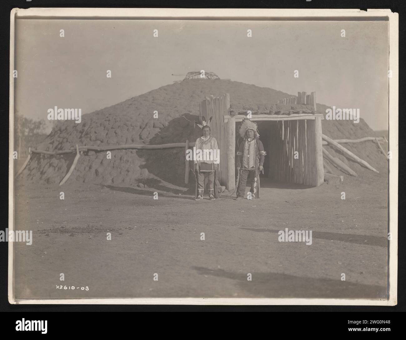 Alfred Chase, 1908. Das Foto zeigt Alfred Chase mit Kopfschmuck und Krähenherz, mit drei Adlerfedern im Haar und einer Halskette mit Bärenkralle, die vor der kuppelförmigen Erdhütte steht. Stockfoto