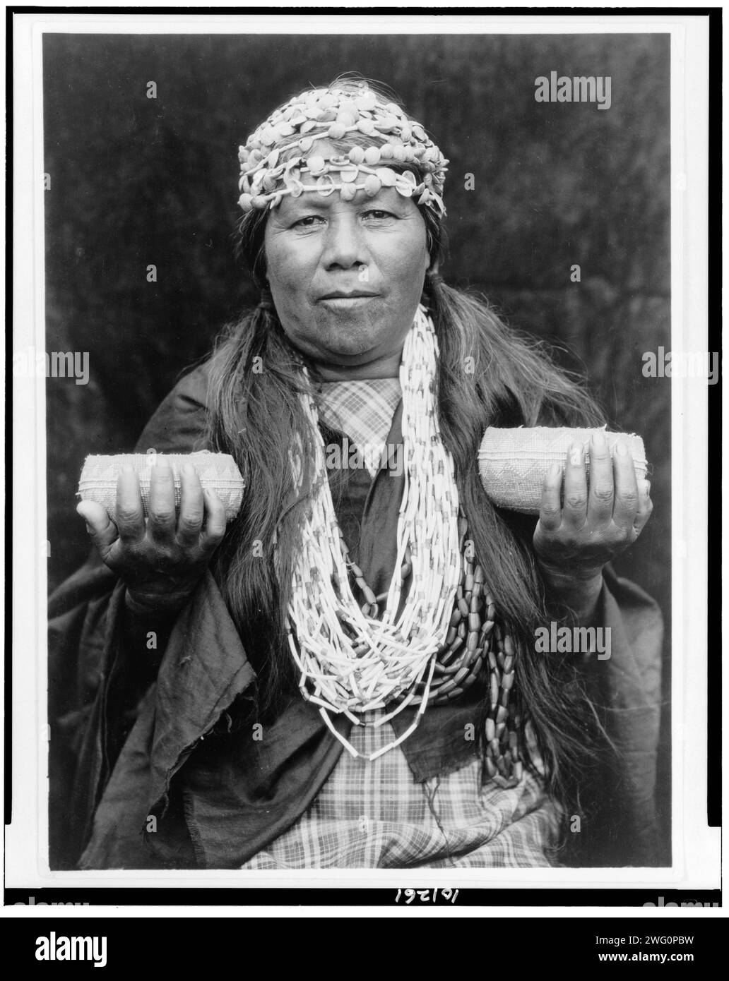 Hupa weibliche Schamane, 1923. Athapascan Hupa Frau aus dem Nordwesten Kaliforniens, halblanges Porträt, stehend, nach vorne gerichtet, mit Muschelkopfbändern, Halskette und zwei Körbe hochhalten. Stockfoto