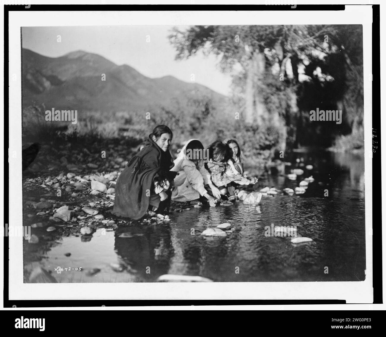 Taos Children, 1905. Vier Taos-Kinder hocken auf Felsen am Ufer des Baches, Berge im Hintergrund. Stockfoto