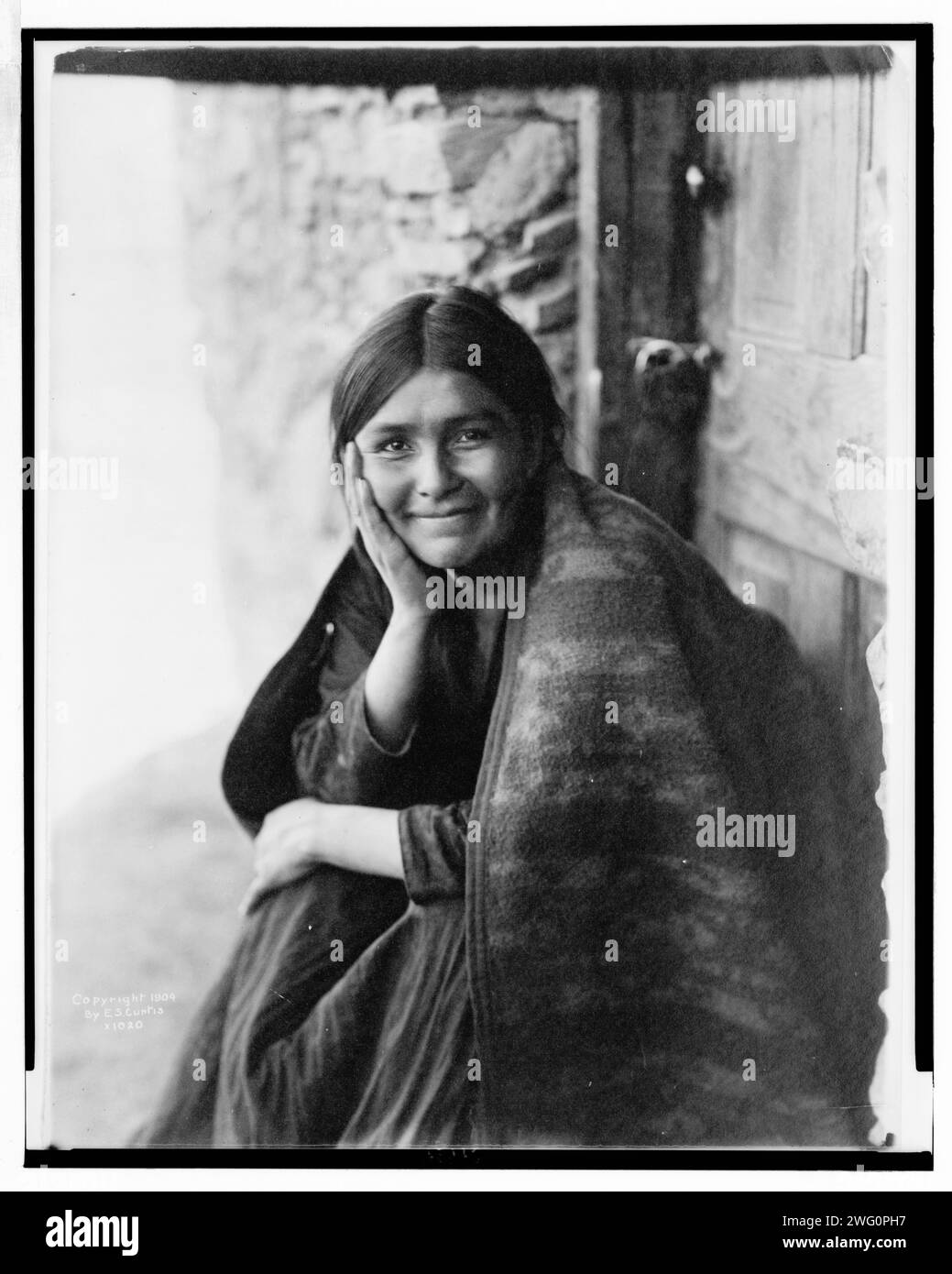 Ein Navaho-Lächeln, 1904. Das Foto zeigt eine Navaho-Frau, die vor der Tür sitzt, mit der rechten Hand auf der Wange, nach vorne gerichtet, lächelnd. Stockfoto