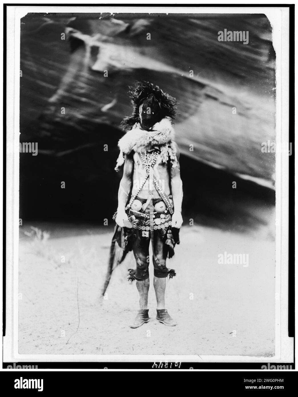 Nayenezgani-Navaho, 1904, 1905. Indisches Porträt in voller Länge, stehend, nach vorne gerichtet, mit dunkler Ledermaske, Pelzkragen, Stoffgürtel, silbernem concho-Gürtel und Ketten. Stockfoto