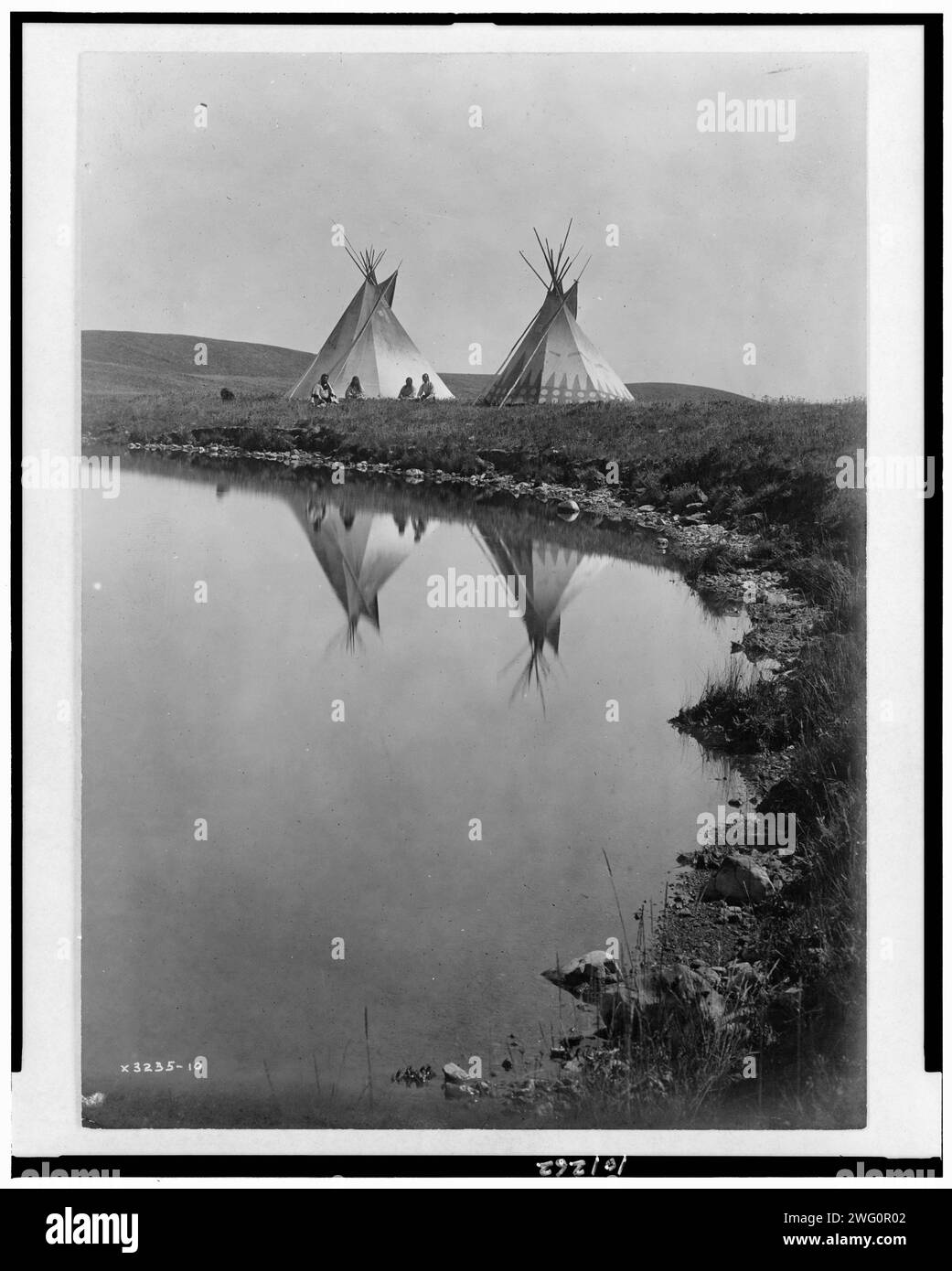 Am Wasserrand - Piegan, 1910. Das Foto zeigt zwei Tipis, die sich im Wasser des Teichs spiegeln, mit vier Piegan-Indianern, die vor einem Tipi sitzen. Stockfoto
