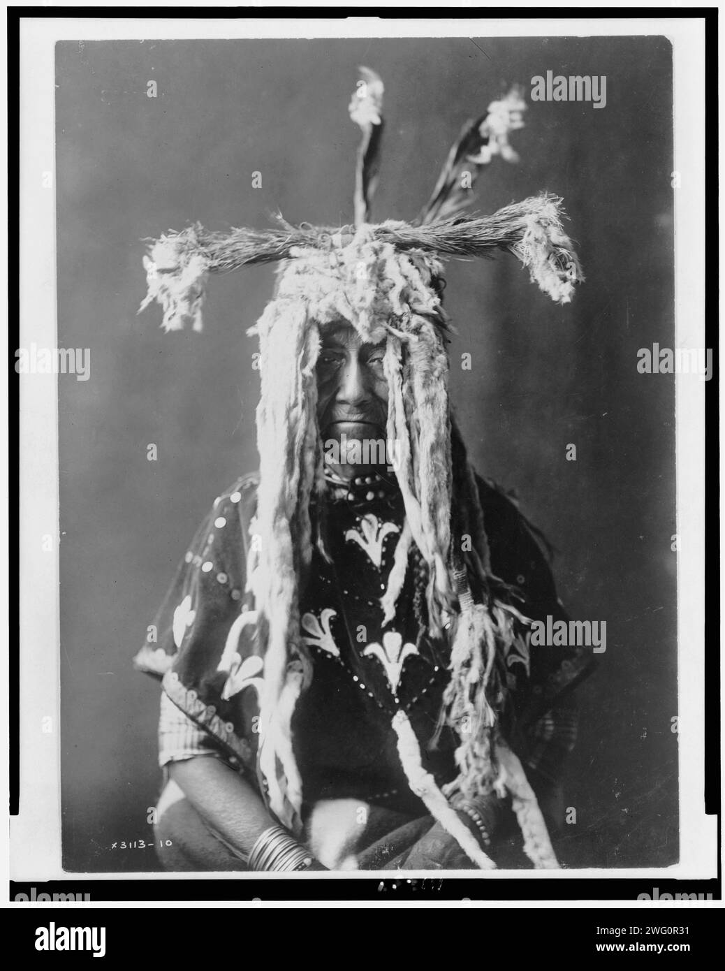 Ersatz-heiliges Kopfkleid-Piegan, 1910. Pieganfrau, halblanges Porträt, sitzend, nach vorne gerichtet, mit Kopfschmuck aus Hermelinschwänzen, Federn und Grassträuben. Stockfoto