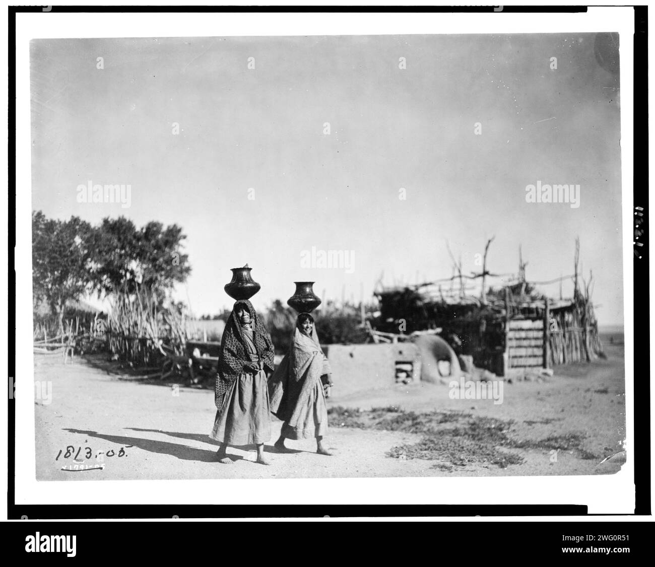 Im Dorf Santa Clara, 1905. Zwei Pueblo-Frauen, die Krüge auf dem Kopf tragen; Bäume, Zugzaun, Stockwerk entlang des unbefestigten Weges im Hintergrund. Stockfoto