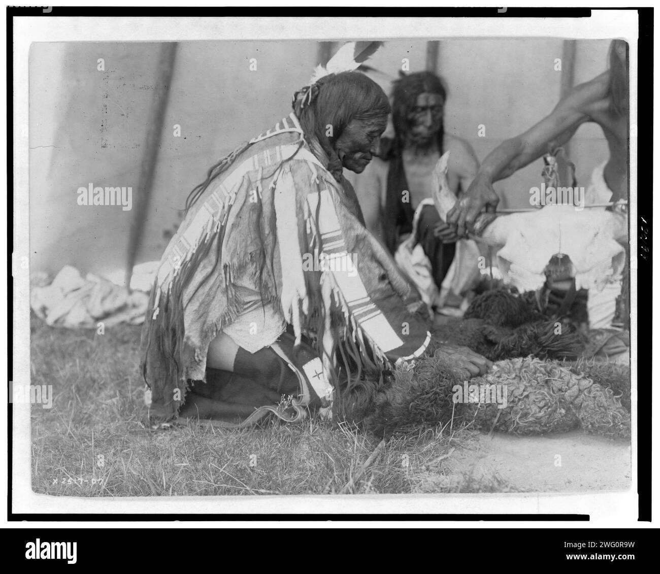 Speichel, Slow Bull (im Hintergrund), Picket Pins Arm, um 1907. Drei Dakota-Männer, sitzend oder kniend auf dem Boden, einer kümmert sich um Bisonschädel. Stockfoto