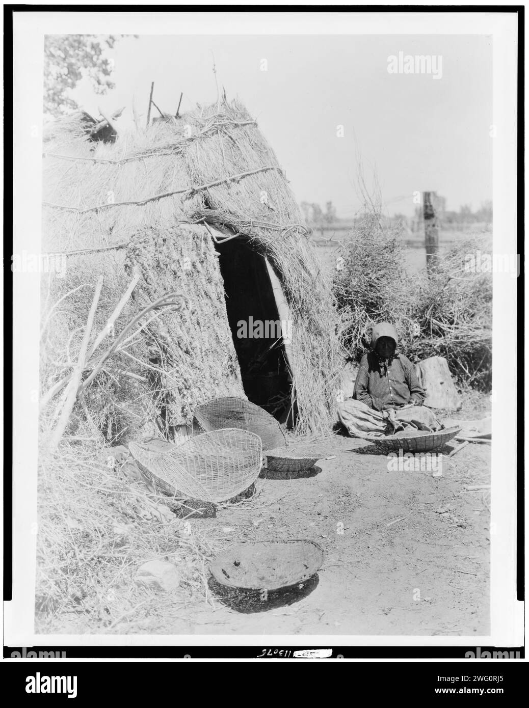 Paviotso-Haus am Walker Lake, 1924. Paviotso-Frau, die vor der strohgedeckten Hütte sitzt, mit Körben, möglicherweise zum Winnowing, im Vordergrund. Stockfoto
