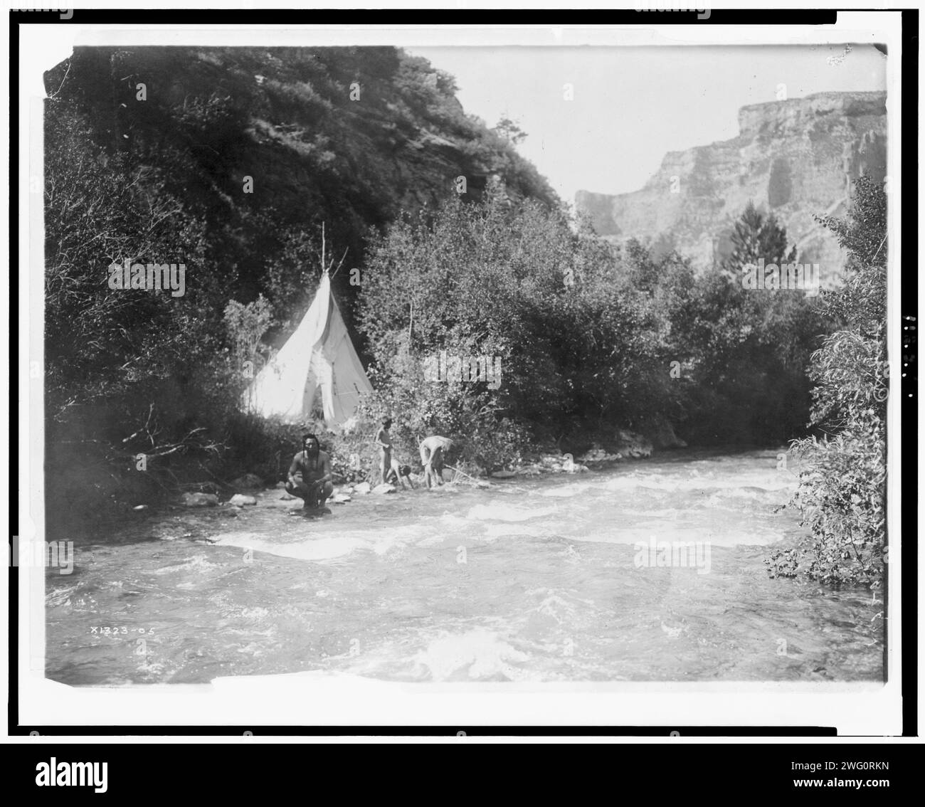 Das Flusslager, 1905. Krähenmann mit zwei Kindern und Frau (?) Am Ufer des Flusses; Tipi, umgeben von Bäumen und Bergen. Stockfoto