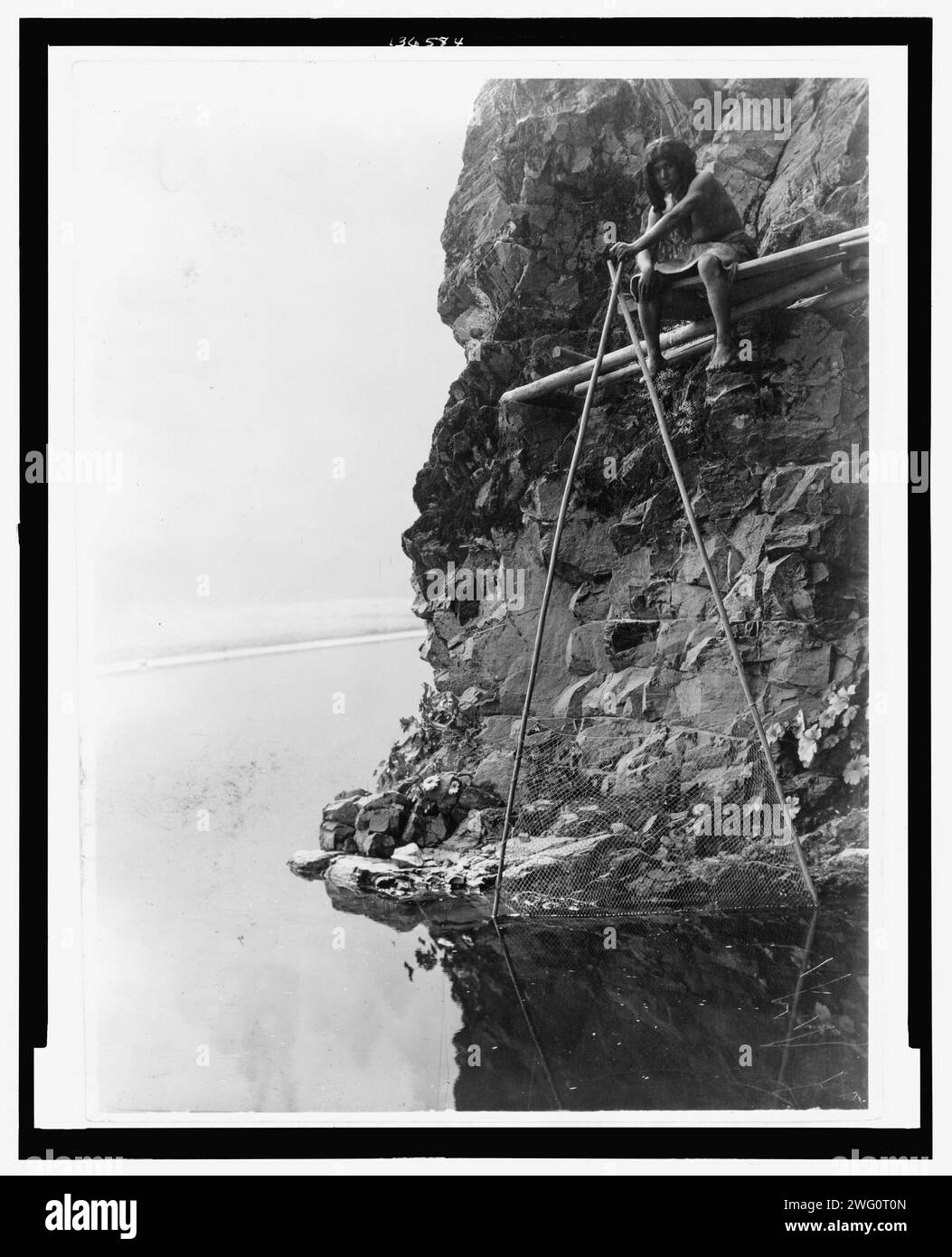 Angelplattform am Trinity River-Hupa, um 1923. Das Foto zeigt einen Hupa-Mann, der auf einer Plattform auf einer felsigen Klippe sitzt und mit einem Fischernetz umgeht. Stockfoto