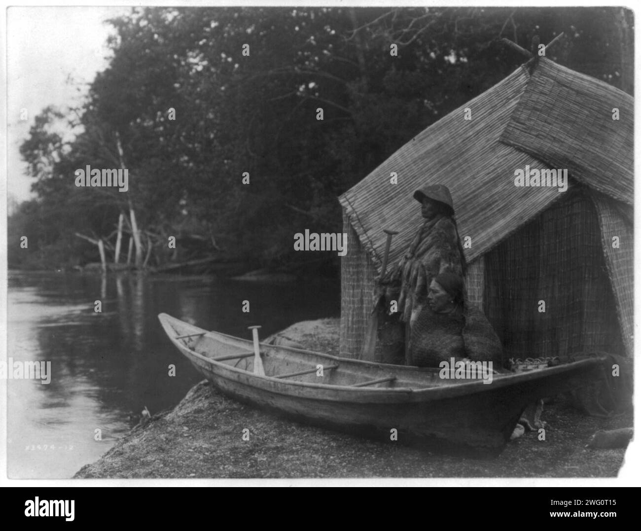 Angellager Skokomish, 1913. Skokomish Paar vor dem Haus aus Schilfmatten, einer sitzt neben dem Kanu, das auf das Ufer gezogen wurde, und der andere steht mit einem Paddel. Stockfoto