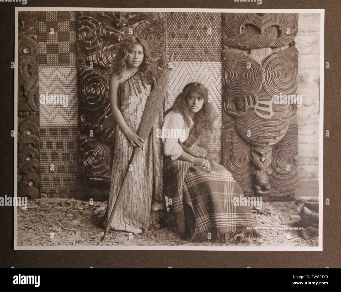 Maori-Mädchen. Hochformat, 1891. Zwei Maori-Mädchen posieren vor einer kunstvoll geschnitzten Innenwand. Beide haben lange Haare und keine Tätowierungen um den Mund. Einer steht und hält ein geschnitztes Holzpaddel. Sie trägt einen steifen Mantel, verziert mit herabhängenden Kordeln, die über eine Schulter um sie gewickelt sind. Der andere sitzt. Sie trägt eine Bluse im europäischen Stil mit einer karierten Wolldecke, die um sie gewickelt ist. Hochformat in voller Länge. Von Charles Appleton Longfellow's Reise nach Neuseeland im Jahr 1891. Stockfoto