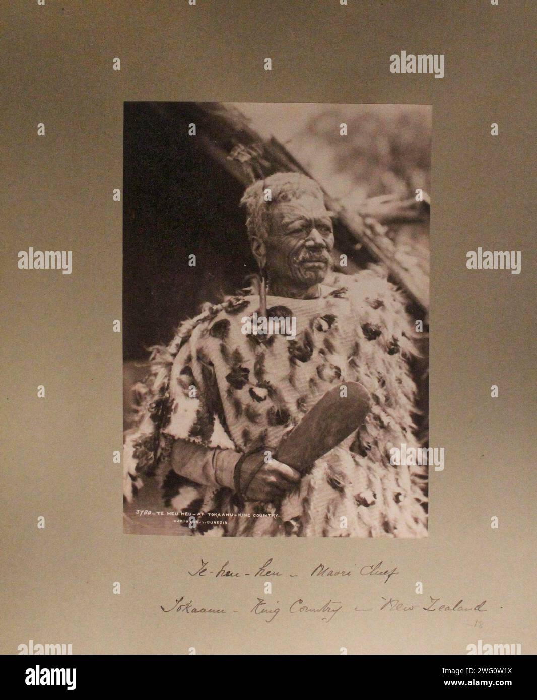 Maori-Häuptling, Te Heu Heu, posiert in der Nähe eines Hauses. Er hat einen Schnurrbart, kurze, weiße Haare, die mit Federn, langen Ohrringen und ausgedehnten Gesichtstattoos verziert sind. Er trägt einen steifen Mantel, der mit Federn um seine Schultern verziert ist. Er hält ein kurzes, hölzernes Paddel in den Händen. Kopf- und Schultern-Porträt. Von Charles Appleton Longfellow's Reise nach Neuseeland im Jahr 1891. Stockfoto