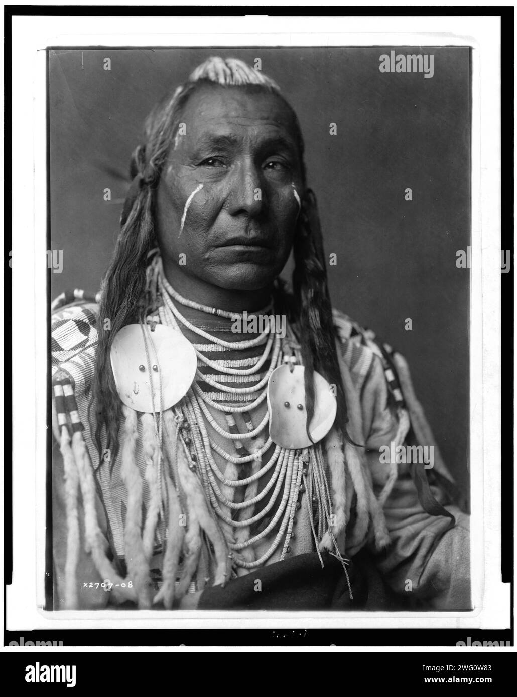 Red Wing-Apsaroke, 1908. Krähen-Indianer, Montana, Kopf-und-Schultern-Porträt, leicht nach rechts gerichtet, weiße Farbstreifen auf Wangen und Haaren, perlenbesetztes Hemd aus Rosinenleder mit Hermelin-Schwänzen oder weißem Kaninchenfell. Stockfoto