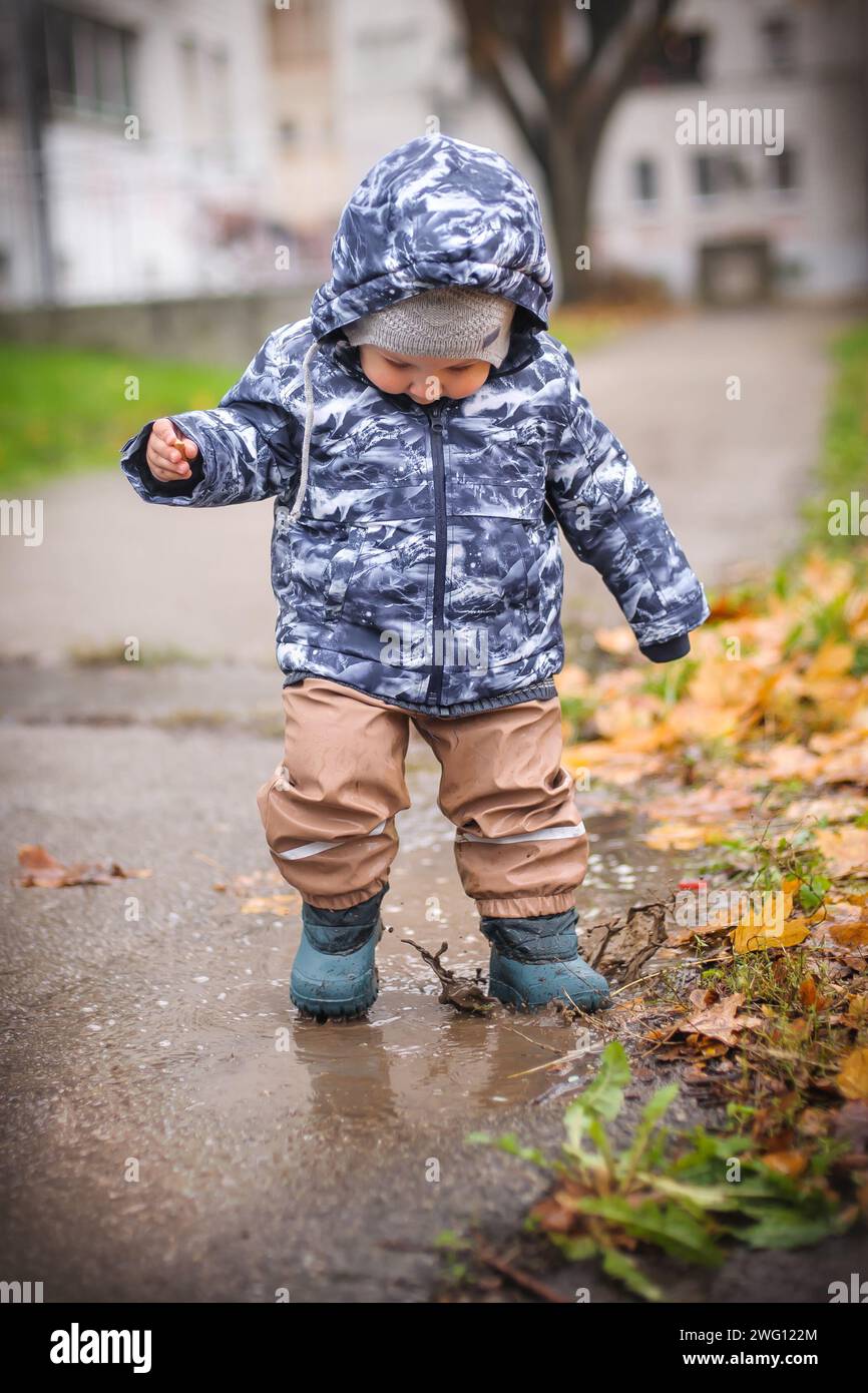 Der kleine Junge steht mitten im strömenden Regen und spielt am regnerischen Tag mit der Regenpfütze auf der Stadtstraße. Abenteuer im Regen Stockfoto