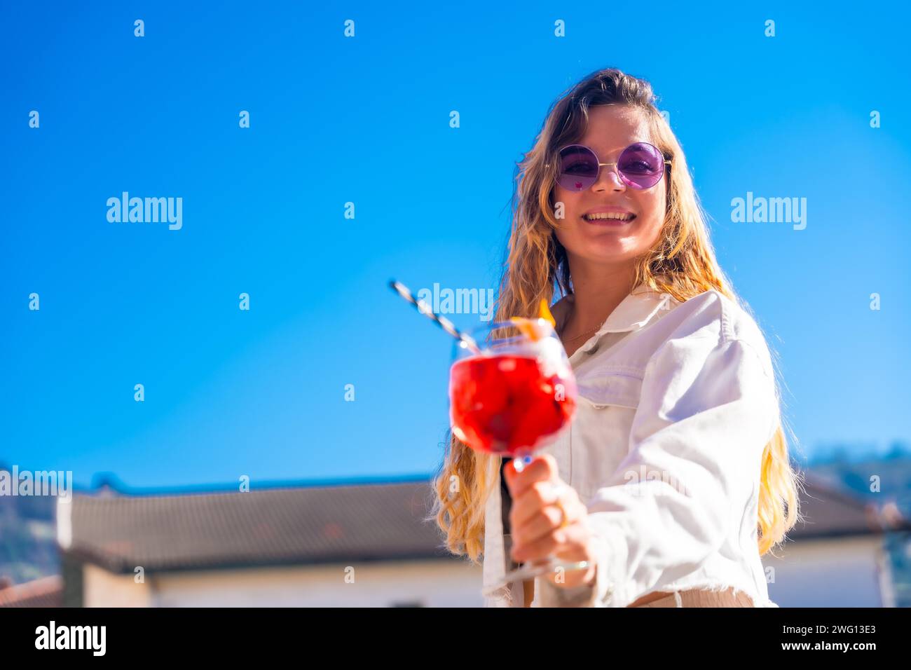 Schicke blonde Frau, die im Sommer mit der Kamera auf dem Dach einen roten Cocktail hält Stockfoto