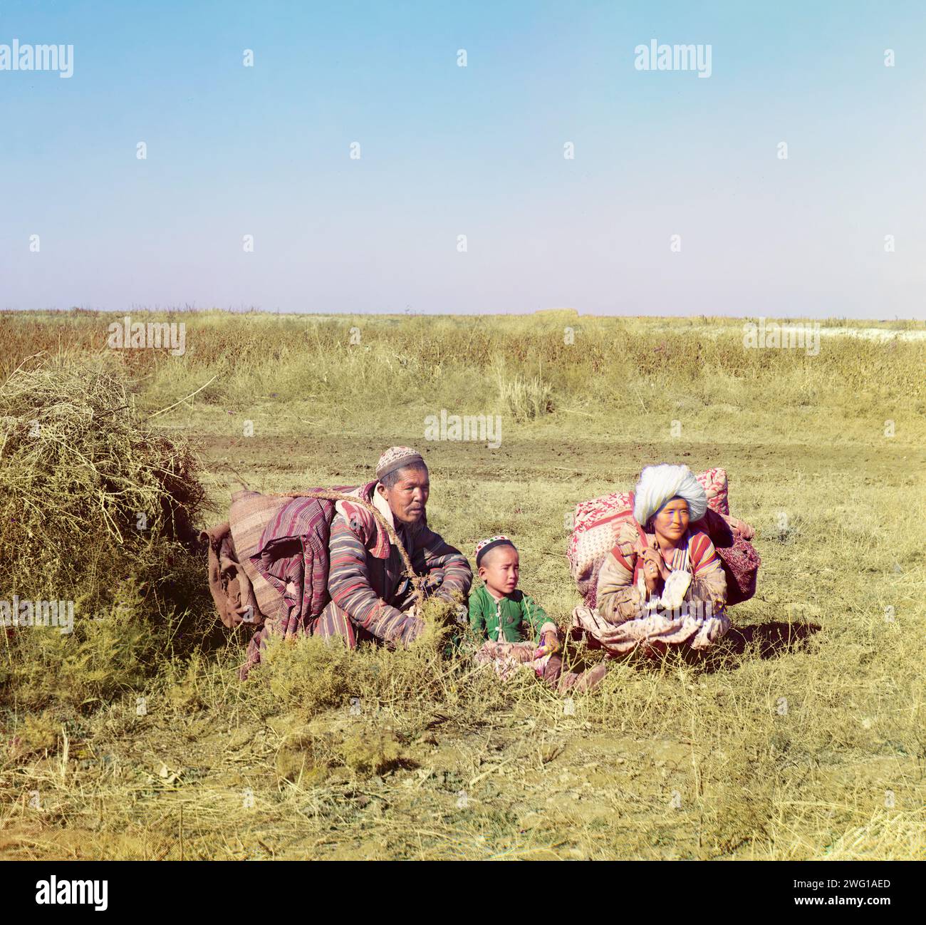 Nomadischer Kirghiz, Golodnaia (oder Golodnaya) Steppe, zwischen 1905 und 1915. Dreiköpfige Familie, kniend auf einem Feld. Viele zentralasiatische Völker, zum Beispiel Kirghiz, Kasachisch und Usbeken, lebten nomadisches Leben auf den Steppen, Tälern und Wüsten und wanderten saisonal von einem Ort zum anderen, da sich die Möglichkeiten für Nahrung, Wasser und Unterkünfte änderten. Hier sehen Sie eine junge kasachische Familie in farbenfroher traditioneller Kleidung, die durch die Steppe Golodnaia (oder „hungrige“) im heutigen Usbekistan und Kasachstan zieht. Stockfoto