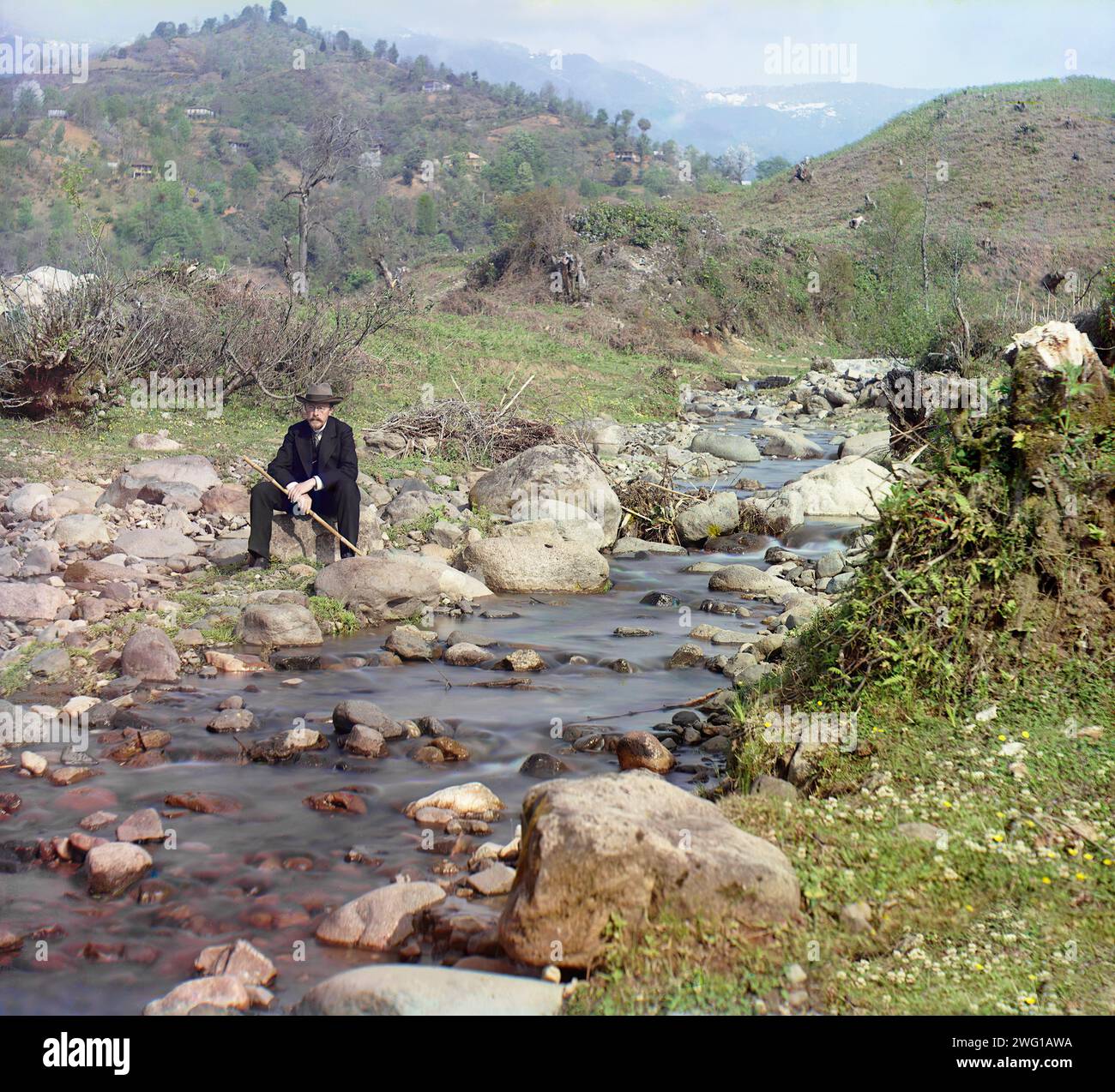 Am Fluss Skuritskhali: Studie, Dorf Orto-Batum, zwischen 1905 und 1915. Der russische Chemiker und Fotograf Sergej Prokudin-Gorski in Anzug und Hut, sitzt auf einem Felsen am Fluss, mit Bergen im Hintergrund, Batumi, Georgien. Stockfoto