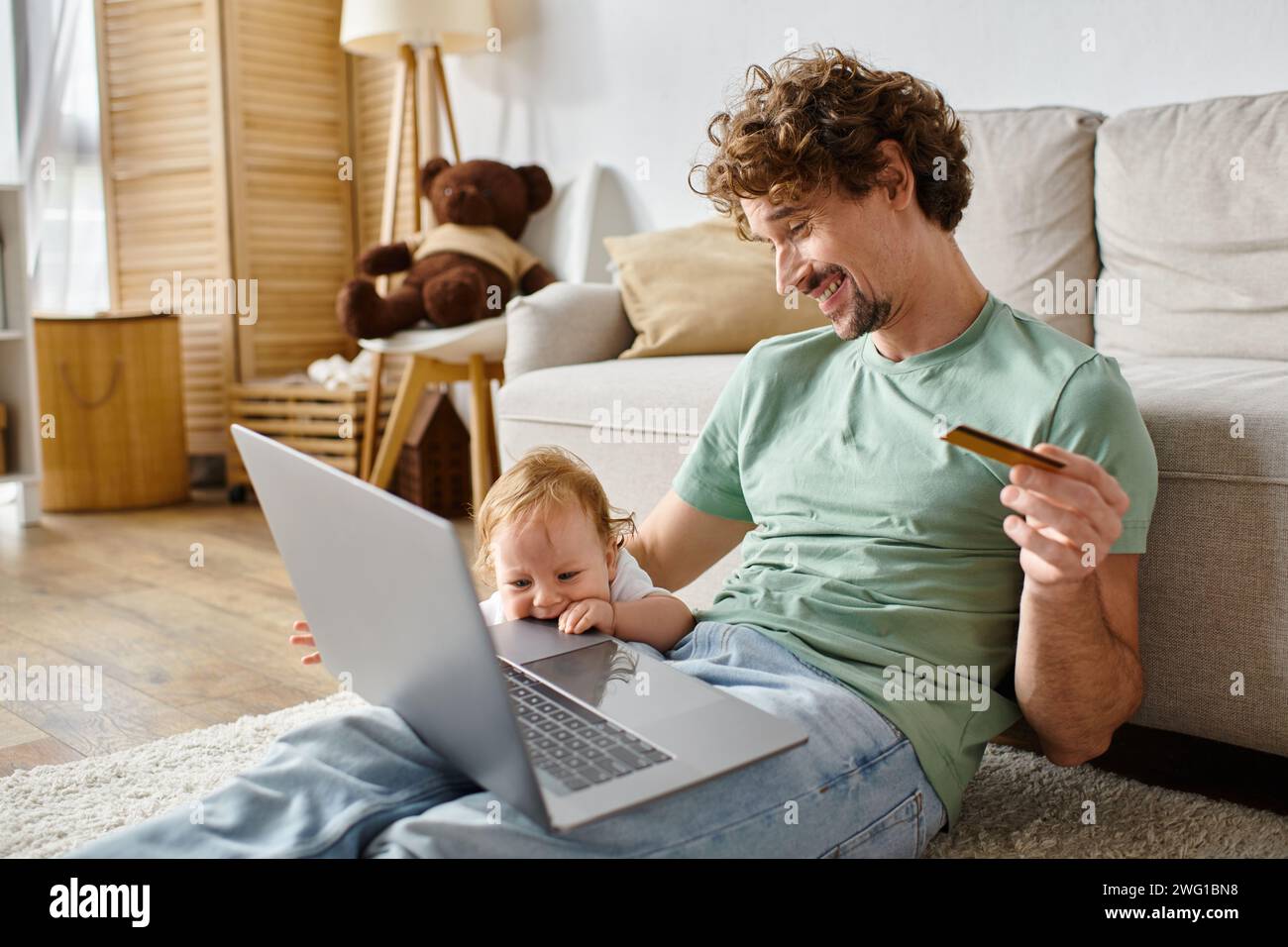 Glücklicher Mann mit lockigen Haaren, der Kreditkarte hält, während er online in der Nähe des kleinen Jungen im Wohnzimmer einkaufen kann Stockfoto