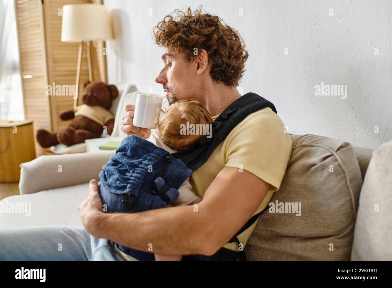 Lockiger Mann mit Säuglingssohn in Babytrage, der eine Tasse Tee im Wohnzimmer hält, Vaterschaft Stockfoto