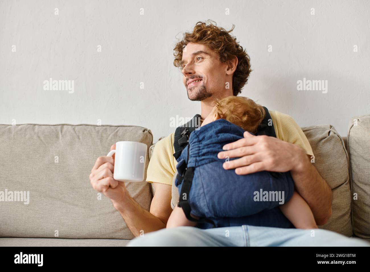 Fröhlicher Vater mit Säuglingssohn in Babytrage, der eine Tasse Tee im Wohnzimmer hält, Vaterschaft Stockfoto