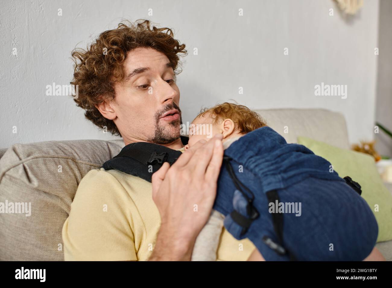 Staunender Mann mit lockigem Haar, der seinen kleinen Sohn in Babytrage, Vaterschaft und Liebe ansieht Stockfoto