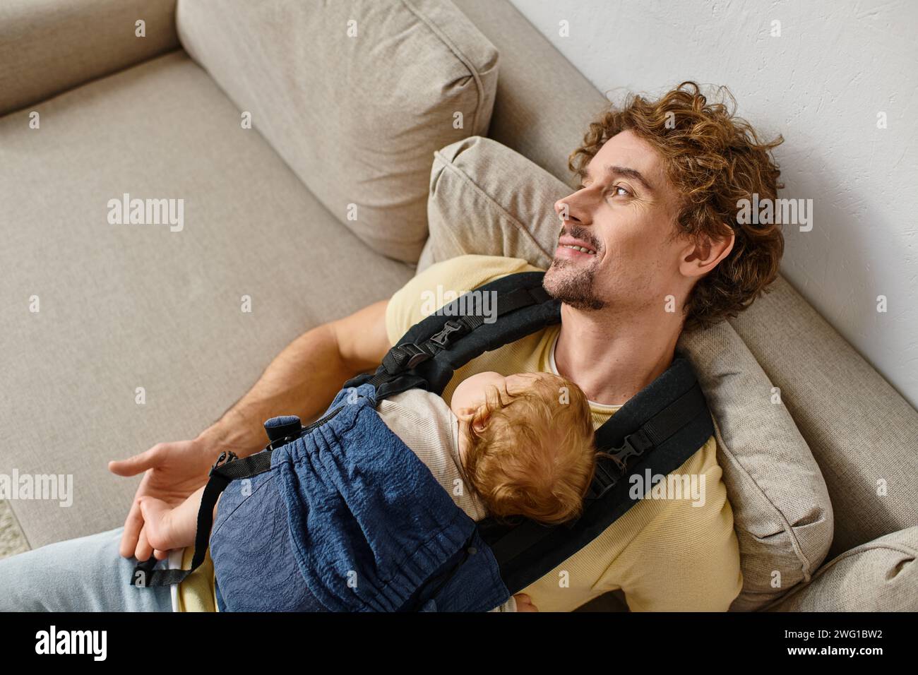 Blick von oben auf den glücklichen Vater, der auf der Couch mit dem Kleinkind in der Babytrage sitzt, Vaterschaft und Liebe Stockfoto