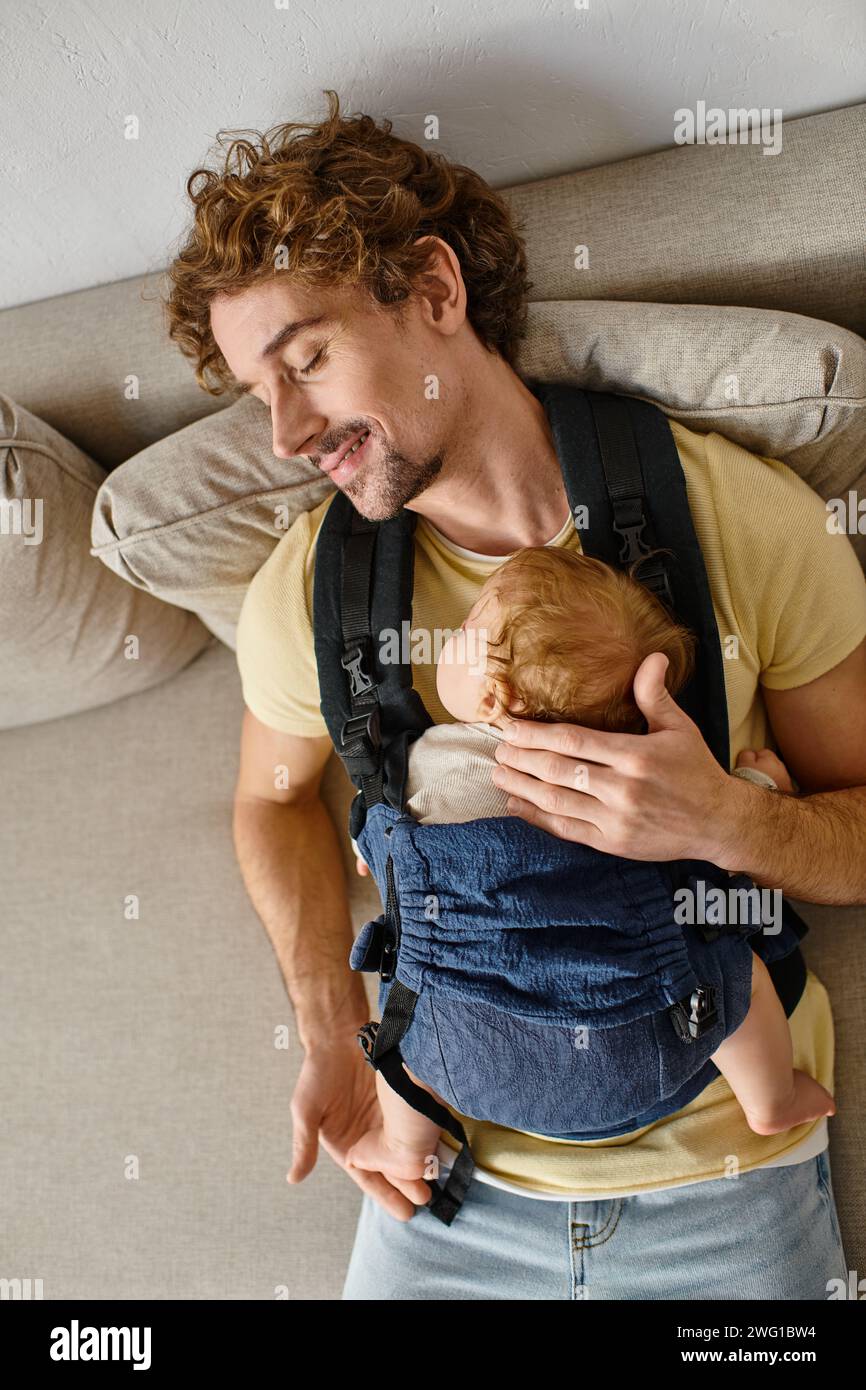 Blick von oben auf den zufriedenen Vater, der auf der Couch mit dem Kleinkind in der Babytrage sitzt, Vaterschaft und Liebe Stockfoto