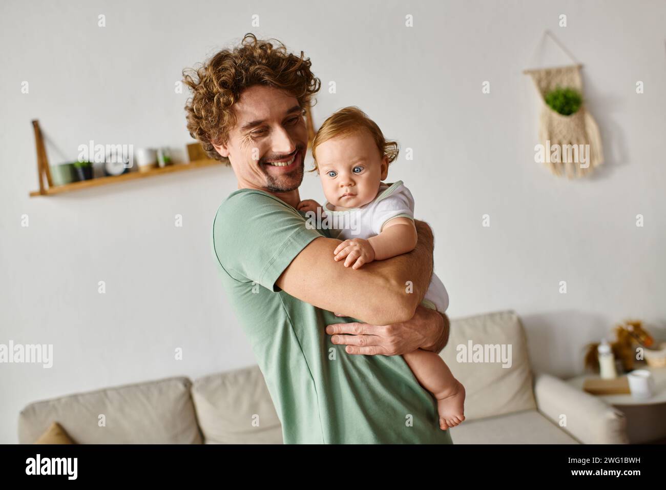 Fröhlicher Vater mit Bart, der seinen kleinen Sohn mit blauen Augen in einem gemütlichen Wohnzimmer hält, Porträt Stockfoto