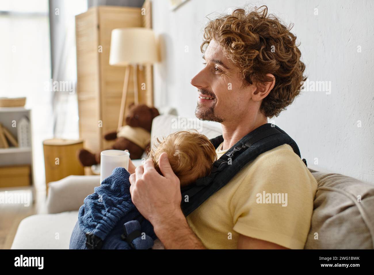 Glücklicher Vater mit Säuglingssohn in Babytrage, der eine Tasse Tee im Wohnzimmer hält, Vaterschaft Stockfoto