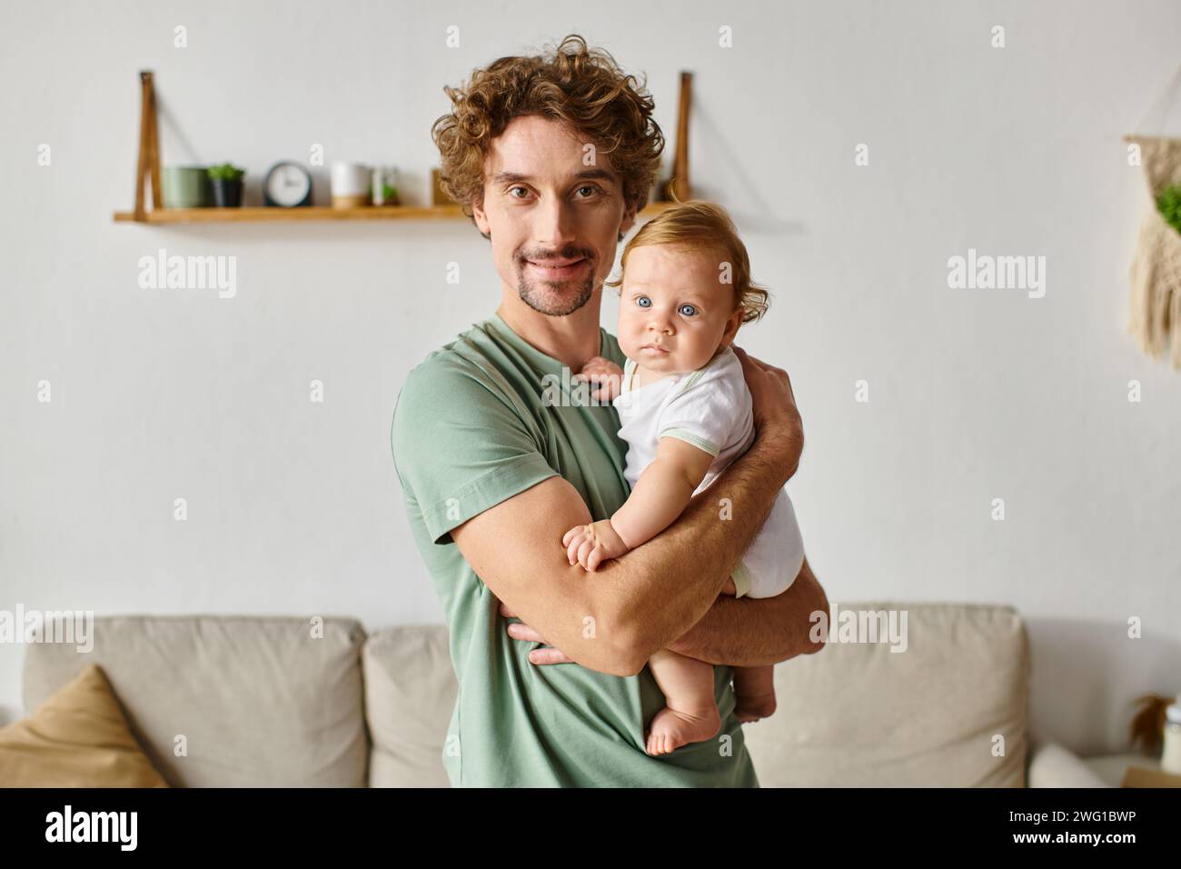 Vater mit lockigen Haaren und Bart, der seinen kleinen Sohn mit blauen Augen in einem gemütlichen Wohnzimmer hält, Porträt Stockfoto
